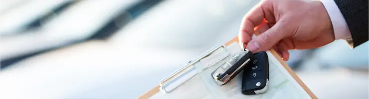 Une main tient des clés de voiture et un presse-papiers avec des documents, devant des voitures neuves.