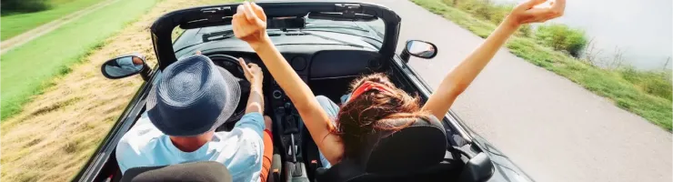 Un couple joyeux sur la route dans une décapotable, les bras levés, longeant un lac.