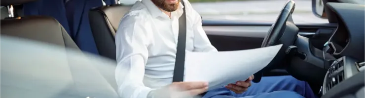 Un homme barbu en chemise lit des documents au volant d'une voiture, un costume à l'arrière.