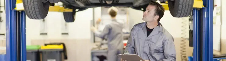 Un mécanicien examine le dessous d'une voiture sur un pont élévateur, avec un presse-papiers.