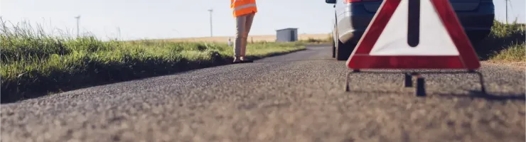 Un triangle de signalisation d'urgence sur la route devant une voiture en panne et une femme.