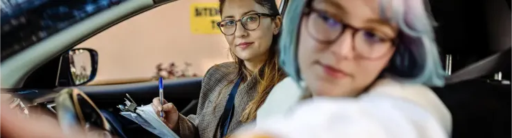 Deux femmes dans une voiture, une conductrice et l'autre tenant un presse-papiers, potentiellement un examen de conduite.
