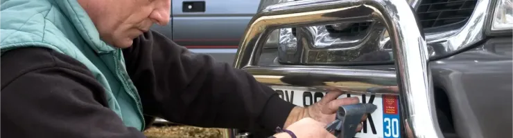 Un homme en veste verte fixe une plaque d'immatriculation sur le pare-buffle avant d'une voiture.