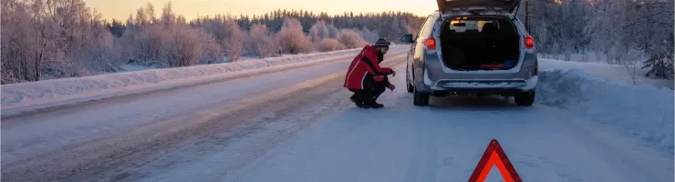 Une personne avec un gilet orange s'accroupit derrière une voiture en panne sur une route enneigée avec un triangle de signalisation.