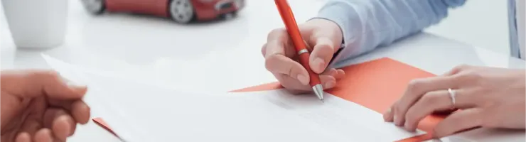 Deux personnes signent des documents d'assurance automobile avec un stylo rouge, une petite voiture rouge est visible en arrière-plan.
