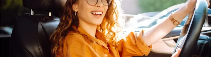 Jeune femme souriante aux cheveux bouclés et lunettes, conduisant une voiture.