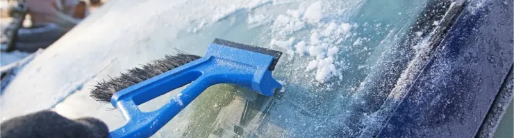 Une main gantée gratte le givre d'un pare-brise de voiture avec un grattoir à neige bleu.