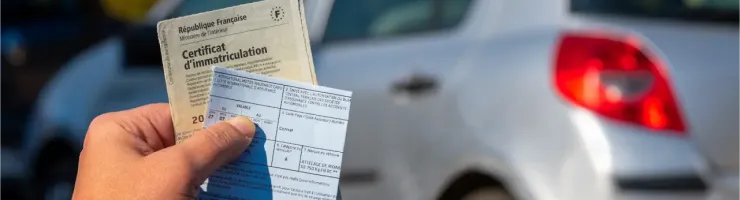 Une main tenant un certificat d'immatriculation et d'autres documents, devant une voiture en arrière-plan.