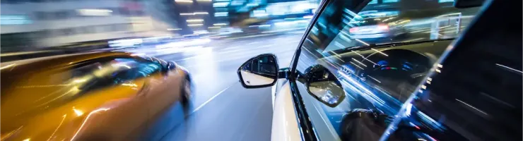 Vue latérale d'une voiture sur une route, avec une autre voiture rapide et les lumières floues de la ville la nuit.