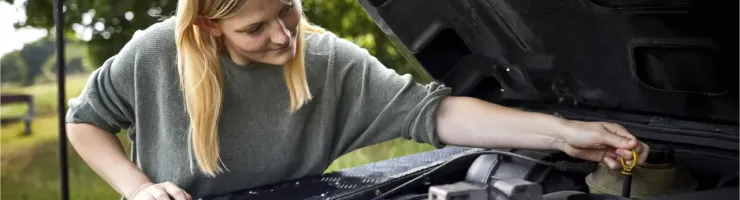 Une jeune femme aux cheveux blonds vérifie le niveau d'huile du moteur d'une voiture, le capot ouvert.