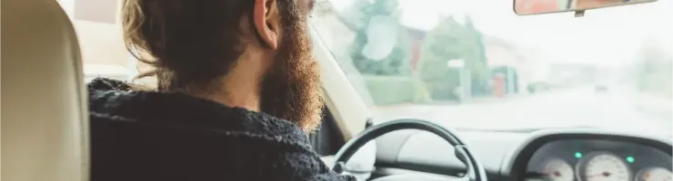 Un homme barbu avec un chignon conduit une voiture, vu de dos, regardant la route devant lui.