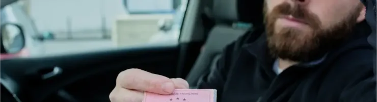 Un homme barbu au regard perplexe tient un permis de conduire français rose vif dans une voiture.