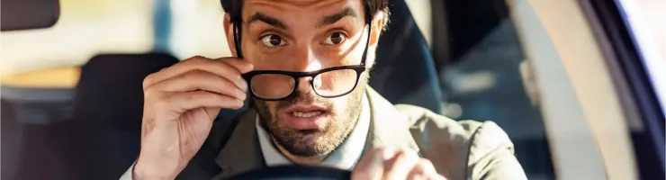 Un homme en costume, l'air surpris, retire ses lunettes en conduisant.