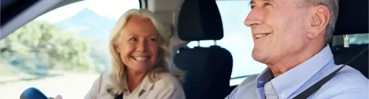 Un couple âgé souriant est assis dans une voiture, l'homme conduit et la femme le regarde.
