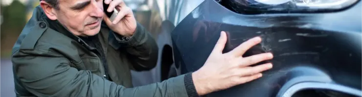 Un homme au téléphone, accroupi à côté de sa voiture accidentée, inspecte des rayures sur le pare-chocs.