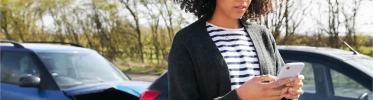 Une femme avec des cheveux bouclés regarde son téléphone, avec deux voitures accidentées en arrière-plan.
