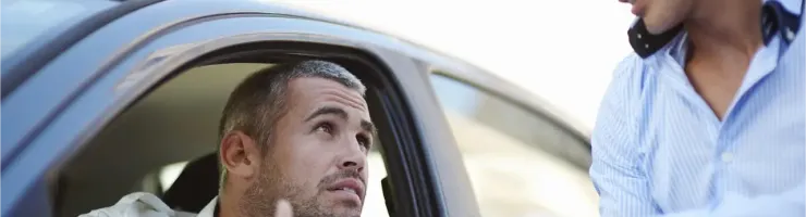 Deux hommes discutent à côté d'une voiture, l'un à l'intérieur, l'autre à l'extérieur.