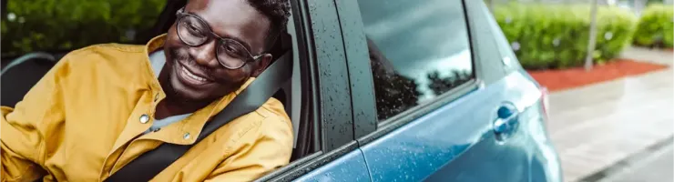 Un homme souriant dans une voiture, le regard tourné vers l'avant, avec des gouttes de pluie sur la carrosserie.