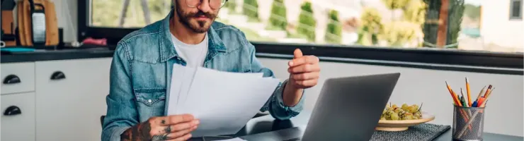 Un homme barbu aux cheveux longs examine des papiers devant son ordinateur portable à table.