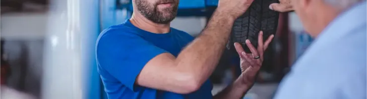 Un mécanicien barbu en blouse bleue explique un pneu à un client dans un garage.