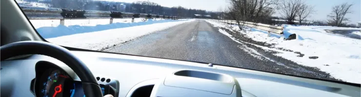 Vue du siège conducteur d'une voiture roulant sur une route enneigée avec des arbres dénudés.
