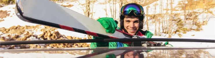 Un skieur souriant attache des skis sur le toit d'une voiture, avec un paysage hivernal en arrière-plan.