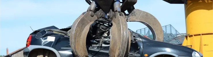 Une grue avec une pince géante soulève une voiture noire fortement endommagée dans une casse.