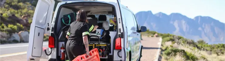 Deux ambulanciers se préparent à sortir leur équipement, dont une planche dorsale orange, d'une ambulance garée au bord d'une route de montagne.