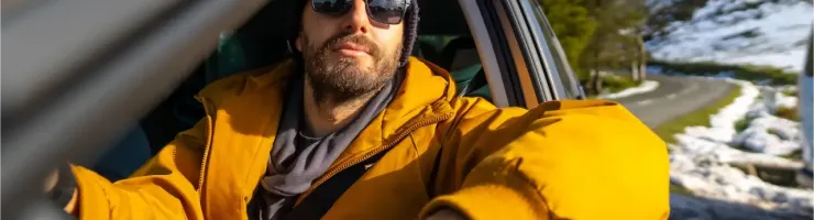 Un homme barbu portant des lunettes de soleil et un bonnet est assis au volant d'une voiture en hiver.