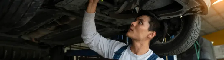 Un mécanicien en salopette examine le dessous d'une voiture sur un pont élévateur, avec un presse-papiers.