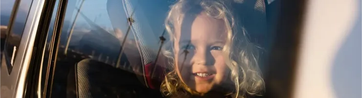 Une petite fille souriante aux cheveux blonds bouclés assise dans un siège auto, vue à travers la fenêtre.