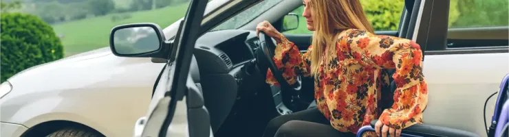 Une femme en robe à motifs floraux sort d'une voiture blanche, une fauteuil roulant à ses côtés.