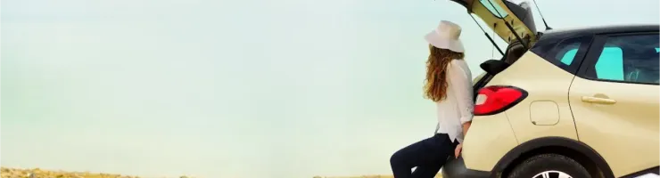 Une femme en chapeau assise sur le coffre ouvert d'une voiture, regardant un lac ou la mer.