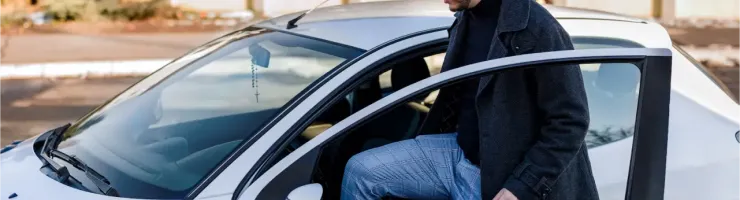 Un jeune homme barbu avec un manteau sombre monte dans une voiture blanche.