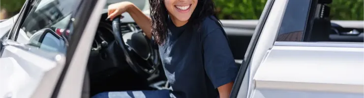 Jeune femme souriante, les cheveux longs et des lunettes, assise sur le siège conducteur d'une voiture.