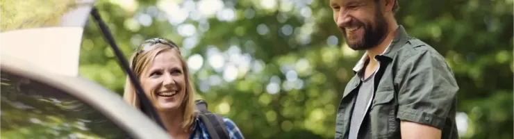 Un couple souriant charge des sacs dans le coffre ouvert d'une voiture, en pleine nature.