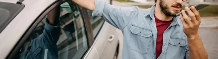 Un homme barbu avec des lunettes et une chemise en jean parle au téléphone près de sa voiture.