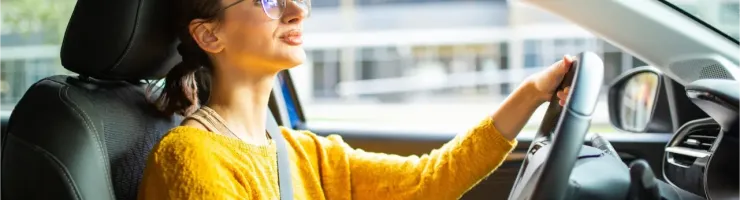 Une jeune femme aux lunettes de vue conduit sa voiture. Elle est vêtue d'un pull jaune vif et semble détendue, appréciant son trajet.