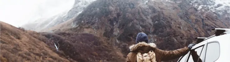 Une personne avec un sac à dos regarde une montagne enneigée près d'une voiture blanche.