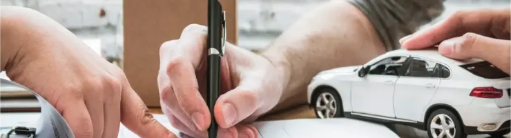 Des mains signent des documents d'assurance auto à côté d'une petite maison et d'une voiture miniature.