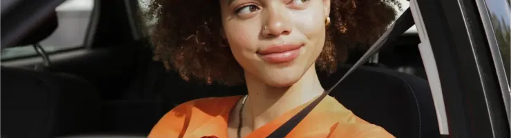 Une jeune femme aux cheveux bouclés sourit en conduisant sa voiture, ceinture bouclée.