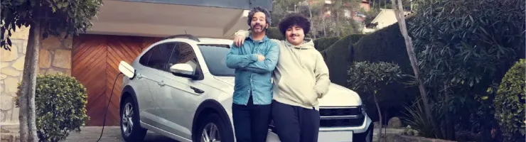 Un père et son fils souriants posent devant une voiture électrique blanche branchée à une borne de recharge devant une maison.