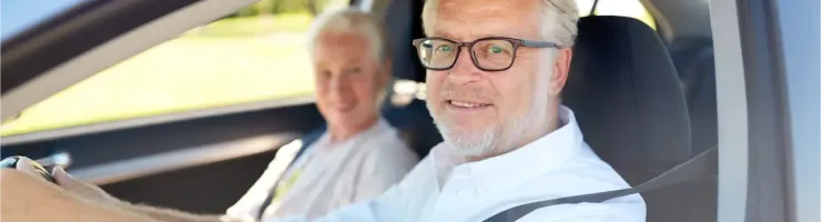 Un homme senior souriant conduit une voiture, sa passagère également souriante à l'arrière-plan.