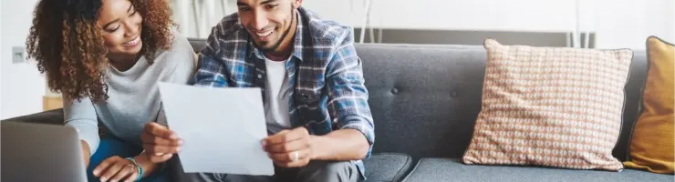 Un couple souriant examine des documents en étant assis sur un canapé avec un ordinateur portable.