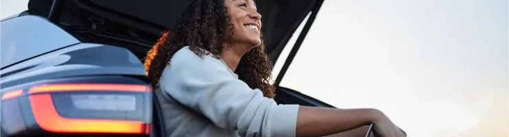 Une jeune femme souriante est assise dans le coffre ouvert d'une voiture avec un ordinateur portable.