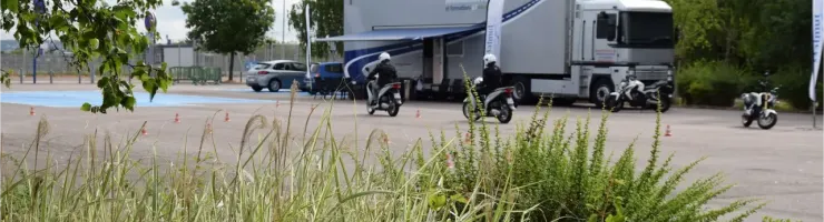 Deux motards sur une piste de formation avec un grand camion d'auto-école en arrière-plan.