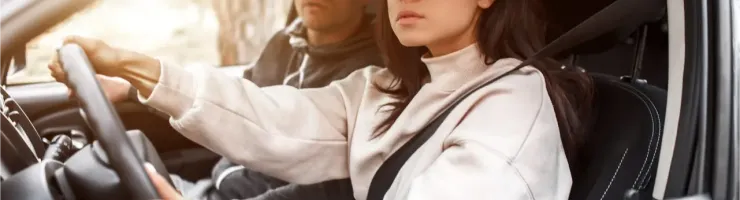 Une femme aux cheveux foncés et lunettes conduit une voiture, regardant attentivement devant elle. Un homme est visible côté passager.