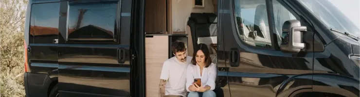 Un jeune couple assis sur le pas de la porte d'un van aménagé noir, utilisant un smartphone par une journée ensoleillée.