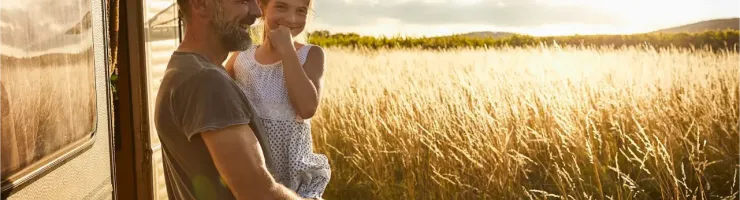 Un père souriant tient sa fille dans ses bras devant un camping-car, avec un champ de blé doré en arrière-plan.