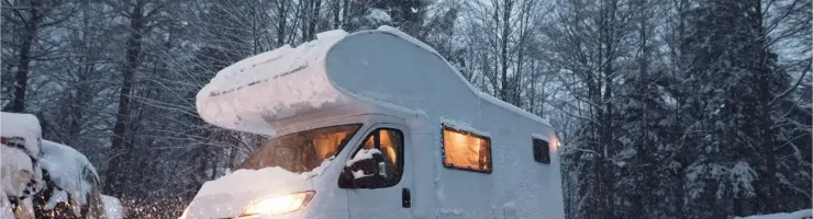 Un camping-car blanc enneigé est garé dans un paysage hivernal boisé, avec les lumières allumées créant une atmosphère chaleureuse.